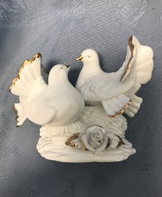 two white doves sitting on top of a vase with roses in the bottom and gold trim