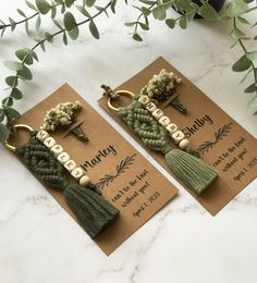 two bookmarks with tassels on them sitting next to some flowers and greenery