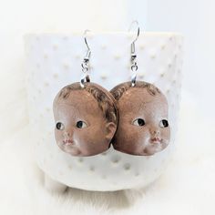 a pair of earrings with an image of a baby's head on it sitting in front of a cup