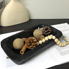 a black tray filled with pine cones and other decorations on top of a table next to papers