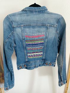 a denim jacket with colorful stitching on the front and back, hanging up against a wall