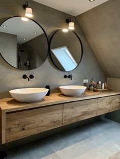 two round mirrors on the wall above two sinks in a bathroom with grey walls and white tile flooring