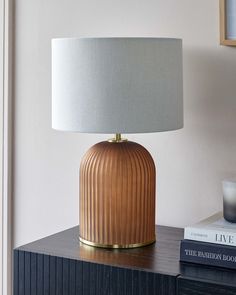 Elegant table lamp with a ribbed bronze base and brass accents, topped with a cylindrical light grey fabric shade, displayed on a black textured side table next to a stack of books. Large Bedside Lamps, Decorating My First Apartment, Brown Table Lamp, Dc Apartment, Colorful Lamp Shades, Virginia House, Earthy Bedroom, Contrast Lighting, Table Lamps Living Room