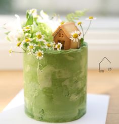 a green cake decorated with daisies and a house in the middle is sitting on a table