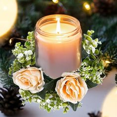 a lit candle surrounded by flowers and greenery on a white table with pine cones