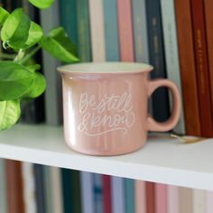 a pink coffee cup sitting on top of a white shelf next to a green plant