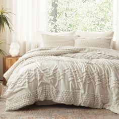 a bed with white comforters and pillows in front of a window, next to a potted plant