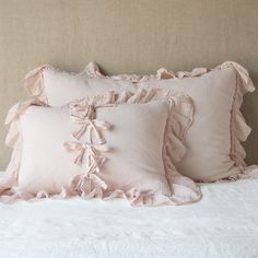 three pink pillows with ruffled edges on a white bedspread in front of a beige headboard