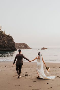 a bride and groom walking on the beach holding hands