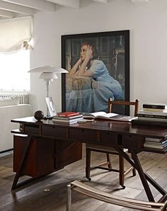 a wooden desk topped with books next to a lamp and painting on the wall behind it