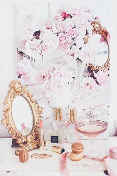 a vanity with pink flowers and cosmetics on it next to a mirror, wine glass and lipstick