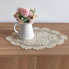 a white vase with pink flowers in it sitting on a wooden table next to a lace doily