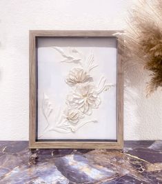 a white flower in a wooden frame sitting on a marble counter next to dried flowers