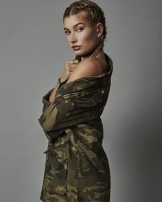 a woman with braids standing in front of a gray background wearing an army jacket