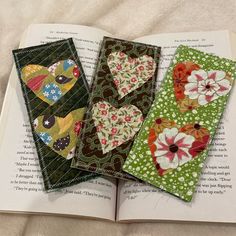 three bookmarks made out of fabric sitting on top of an open book with the pages folded down