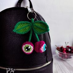 a crocheted cherry and leaf decoration on a black leather backpack next to a bowl of cherries