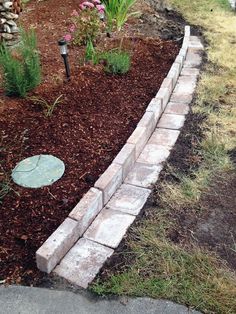 a brick garden border with mulch grass and flowers in the back ground, next to a stone path