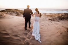 a bride and groom walking through the sand dunes at sunset on their beach wedding day