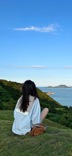 a woman sitting on top of a lush green hillside next to the ocean with a sky background