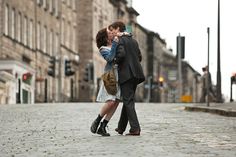 a man and woman kissing on the street in front of an old building with cobblestones