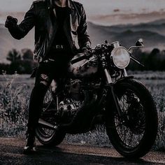 a man sitting on top of a motorcycle in the middle of a field with mountains behind him