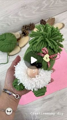 a hand holding a green stuffed animal on top of a pink piece of paper with pine cones around it