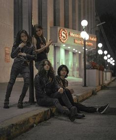 four young women sitting on the curb in front of a building at night with their arms around each other