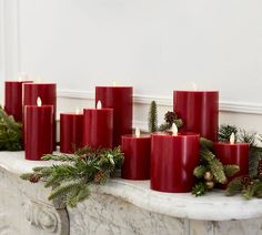 red candles are lined up on a mantle with evergreen branches and pine cones around them