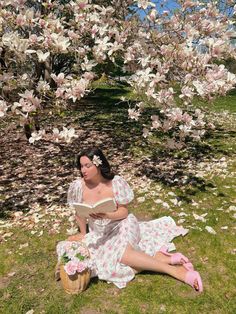 a woman sitting on the ground reading a book in front of a tree filled with flowers