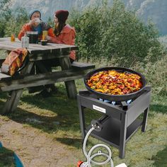 two people sitting at a picnic table with a large grill on the ground next to them