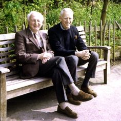 two older men sitting on a wooden bench in the park, one is smiling at the camera