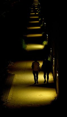 two people walking down a dark street at night with their shadows on the ground and one person holding hands