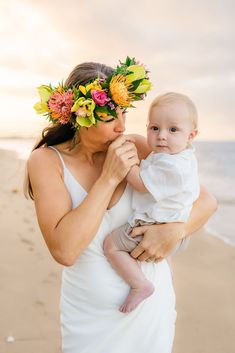 a woman in a white dress holding a baby on the beach with flowers in her hair