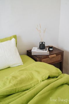 a bed with green sheets and pillows in a small room next to a nightstand with a book on it