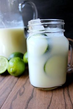 two glasses filled with liquid next to limes on a wooden table and one has a green drink in it