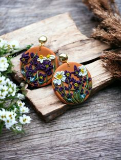 two orange earrings with purple and white flowers on them sitting next to some daisies