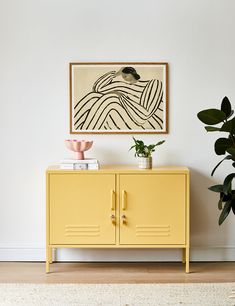 a yellow cabinet sitting in front of a painting on the wall next to a potted plant