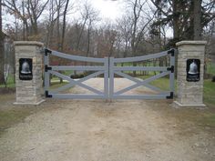 an open gate with two bells on it in the middle of a dirt road surrounded by trees