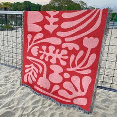 a red and white blanket sitting on top of a sandy beach next to a fence