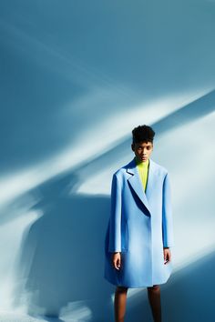 a man in a blue coat and yellow shirt standing against a white wall with his hands on his hips