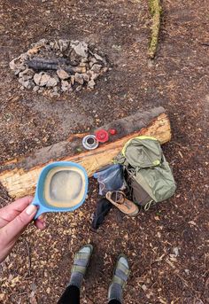 a person holding a blue cup in front of a campfire and camping gear on the ground