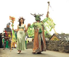 two people dressed in costume walking down a street with other people behind them and one person holding a staff