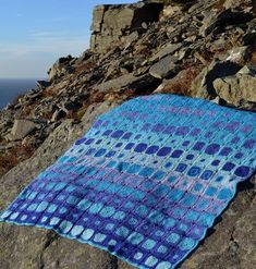 a blue and white crocheted blanket sitting on top of a rocky hill next to the ocean