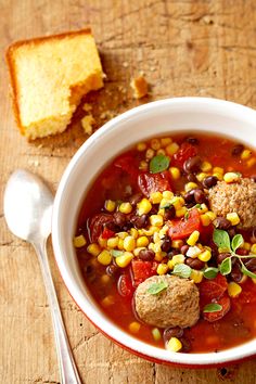 a bowl of soup with meatballs, tomatoes and corn on the side next to a piece of bread