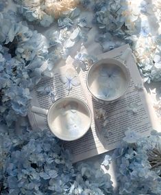 two cups and saucers on top of an open book surrounded by blue hydrangeas