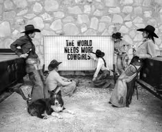 black and white photograph of people with dogs in front of a sign that says the world needs more cowgirls