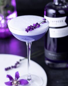 a white plate topped with a purple drink next to a bottle of alcohol and flowers