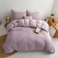 a bed with purple comforter and pillows in a white room next to a wooden table