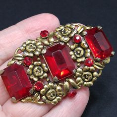 *Description: This is a beautiful floral Art Nouveau emerald cut red glass brooch with gold flowers on a wide brooch from the 1930s to 1940s. The gold flowers flow between the six red rhinestone chatons and three evenly placed large emerald cut red glass rhinestones. There is an early rolling c clasp and long pin on the back reaching from one side to the other. The large stones are open back, allowing the light to shine through. The metal is gold toned pot metal. This would be a great addition t Red Formal Costume Jewelry Brooches, Vintage Red Brooches For Formal Occasions, Vintage Red Brooches For Party, Vintage Red Wedding Brooches, Antique Red Wedding Brooch, Antique Red Brooch As Gift, Red Rectangular Art Deco Jewelry, Antique Red Brooch Jewelry, Retro Red Brooch Jewelry