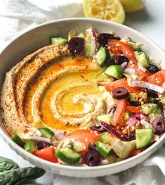 a white bowl filled with hummus, olives and other vegetables next to a lemon wedge